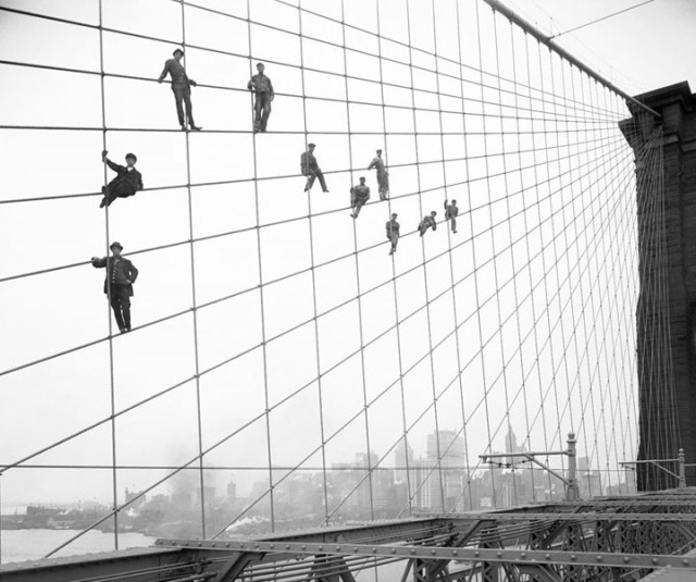 Photo ancienne en noir et blanc de plusieurs ouvriers debout à plusieurs mettre de hauteur sur les cables qui relient les colonnes du pont de Brooklyn au sol.