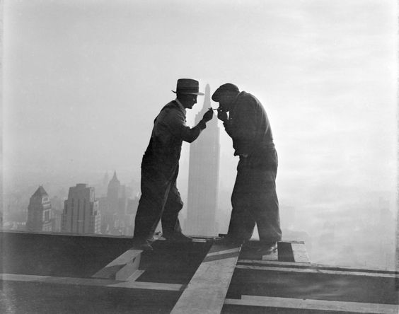 Photo ancienne en noir et blanc de deux ouvriers debout sur une charpente en acier à plusieurs dizaines de mètres de hauteur. Celui de gauche au chapeau élégant, allume la cigarette du second ouvrier à droite.